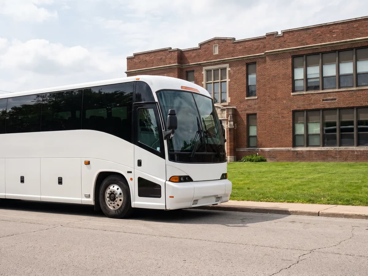 A white coach bus parked along a curb beside a red-brick school building and a green lawn