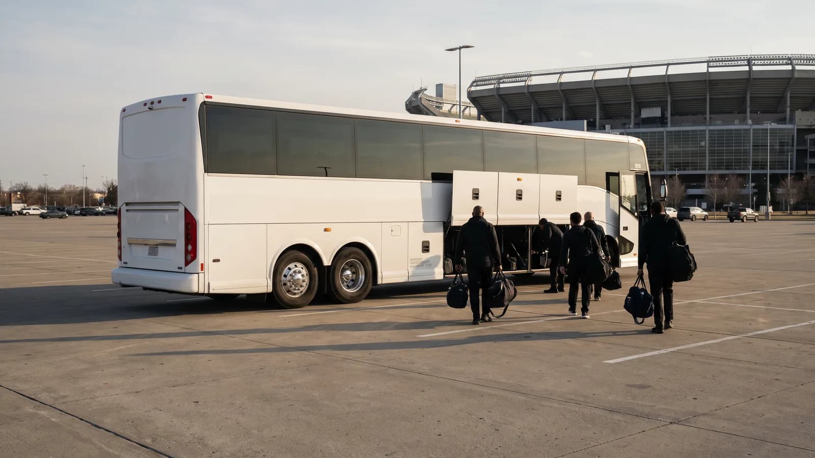 Adult players carrying equipment bags to a white coach bus in a stadium parking lot